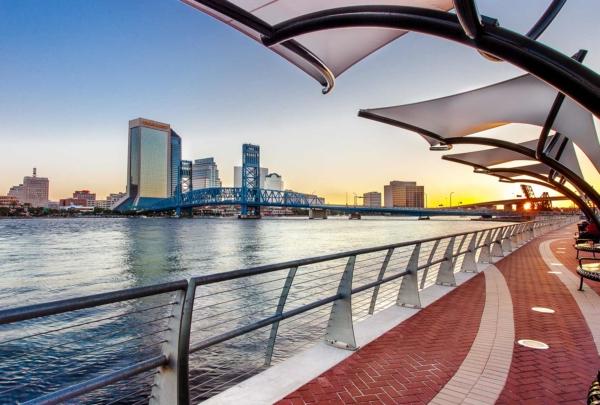 Photo of Jacksonville's 南岸河边漫步 at sunset. Red brick sidewalk alongside the river with steel guardrails. 天篷悬在人行道上. 背景是杰克逊维尔的天际线.